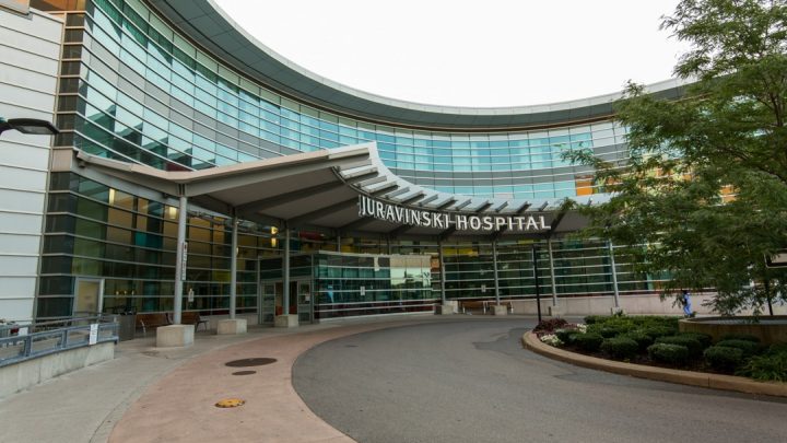 The front entrance of the Jurvanski Hospital during the day that is covered in floor to ceiling windows. In front of the doors is a space for cars to drop people off at the doors.