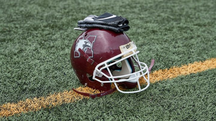A maroon football helmet with the McMaster marauders symbol on the side, resting on a football field. Resting on top of the helmet is a set of football gloves.